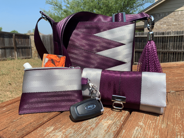 Purse, pouch and wallet made from purple and silver seat belts sit on a wooden table.