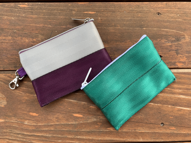 Two zippered seat belt pouches sitting on a wooden surface. One is silver and purple, the other is green